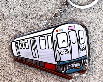 Puede incluir: Un llavero de tren blanco y gris con un diseño de cara sonriente. El llavero tiene un anillo y una cadena de metal plateado. El tren tiene detalles en rojo, azul y rosa. El tren está sobre una vía marrón.