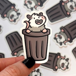 May include: A collection of stickers featuring a cartoon opossum in a trash can. The opossum is gray and white with pink ears and a pink tongue. The trash cans are dark gray with a brown rim.