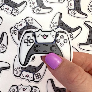 May include: A white and black video game controller sticker with a cute cartoon cat face on the front. The controller has buttons and a joystick. The sticker is on a white background.