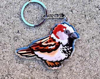 May include: A colorful bird-shaped keychain. The keychain features a detailed illustration of a sparrow with brown, white, red, and black markings. It has a silver-colored keyring and chain. The bird is perched on a gray surface.