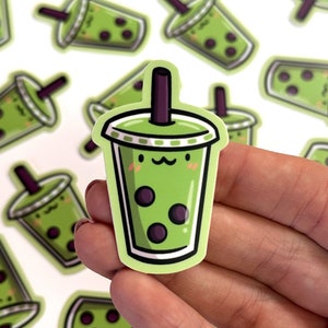 May include: A green and white sticker of a cartoon boba tea cup with a smiling face and black boba pearls. The sticker is on a white background.