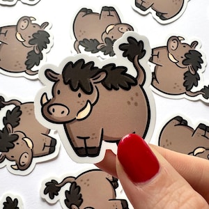 Könnte beinhalten: Eine Sammlung von Cartoon-Warzenschwein-Aufklebern. Die Aufkleber zeigen ein braunes Warzenschwein mit dunkelbraunem Fell, Stoßzähnen und einem lockigen Schwanz. Die Aufkleber befinden sich auf weißem Hintergrund und werden von einer Hand mit einem rot lackierten Nagel gehalten.