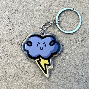 Może przedstawiać: Brelok w kształcie fioletowej chmury z kreskówki z błyskawicą. Chmura ma zły wyraz twarzy z zmarszczonymi brwiami i brwiami.
