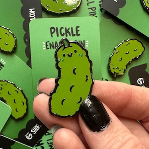 May include: A green pickle enamel pin with a smiling face and a black outline. The pin is on a green background with the text "PICKLE ENAMEL PIN" in white.