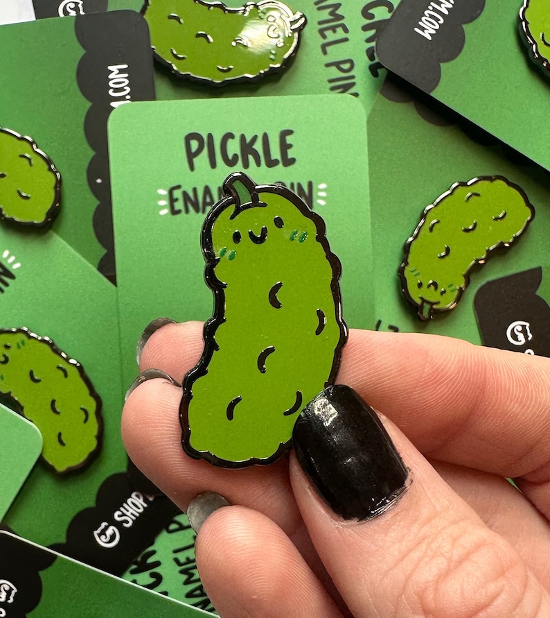 May include: A green pickle enamel pin with a smiling face and a black outline. The pin is on a green background with the text "PICKLE ENAMEL PIN" in white.