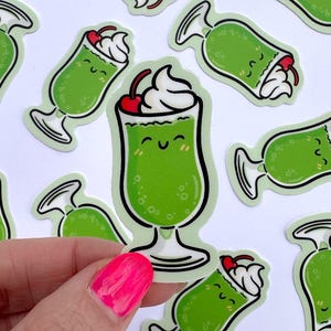 Puede incluir: Un conjunto de pegatinas de dibujos animados verdes y blancas con un batido verde sonriente con una cereza en la parte superior. Las pegatinas están sobre un fondo blanco.