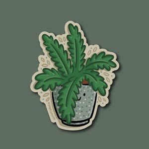 May include: A sticker featuring a cartoon illustration of a potted plant. The plant has large, green, fern-like leaves and is in a gray pot with white polka dots. The sticker has a white border and a green background.