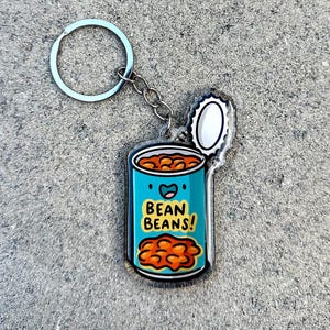 May include: A blue and yellow bean can keychain with a smiling face and the text "BEAN BEANS!" The can is open and the lid is attached to the keychain.
