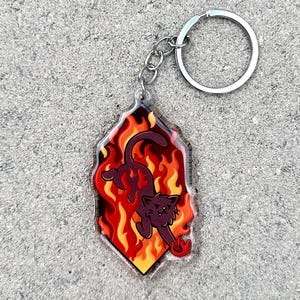 Puede incluir: Un llavero de gato negro con un fondo de acrílico transparente. El gato está representado en un diseño de llamas naranja y amarillas.