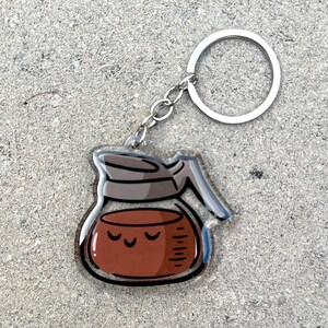 May include: A clear acrylic keychain shaped like a coffee pot with a brown lid and brown coffee inside. The coffee has a cartoon face with three closed eyes.