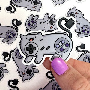 Op de afbeelding: Een set grijze en witte katten stickers met een ontwerp van een videogamecontroller. De stickers tonen een kat die een controller vasthoudt met knoppen en een snoer.