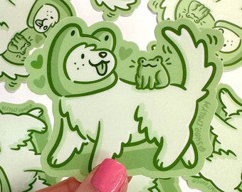 Puede incluir: Pegatina verde y blanca con un perro de dibujos animados que lleva un sombrero de rana verde y una rana verde sentada en su espalda. La pegatina tiene el texto "@shop_emily_m" en la esquina inferior derecha.
