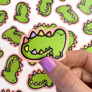 Op de afbeelding: Een set kleurrijke dinosaurus stickers met roze, groene en paarse accenten. De stickers tonen een cartoon dinosaurus met een vriendelijke uitdrukking.