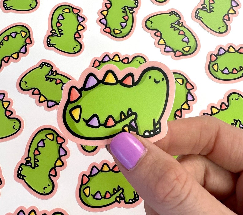 Op de afbeelding: Een set kleurrijke dinosaurus stickers met roze, groene en paarse accenten. De stickers tonen een cartoon dinosaurus met een vriendelijke uitdrukking.