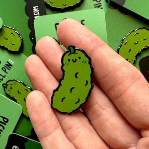 May include: A green enamel pin shaped like a pickle with a smiling face and a black outline. The pickle has a small stem on top and is slightly curved.
