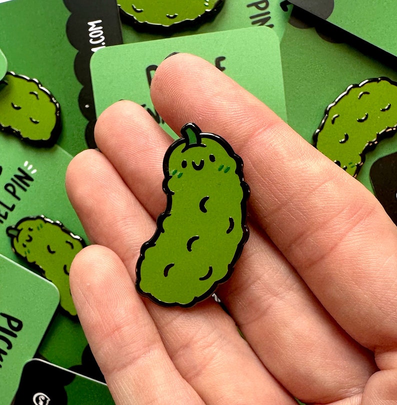 May include: A green enamel pin shaped like a pickle with a smiling face and a black outline. The pickle has a small stem on top and is slightly curved.
