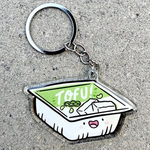 May include: A clear acrylic keychain shaped like a tofu container with a smiling face. The container has green edgings and the text "TOFU!" printed on the top. There are green edamame beans inside the container.