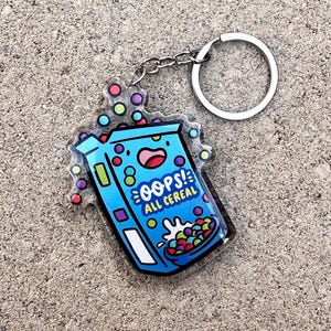 May include: A blue acrylic keychain featuring a cartoon cereal box design with the text "OOPS! ALL CEREAL". The keychain has a silver keyring and chain, with colorful bubble accents.