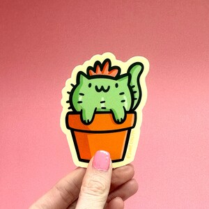 May include: A green cat-shaped cactus with a red flower on top, sitting in an orange pot. The sticker has a yellow outline and the text "@juneberryart" at the bottom.