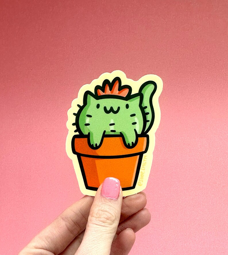 May include: A green cat-shaped cactus with a red flower on top, sitting in an orange pot. The sticker has a yellow outline and the text "@juneberryart" at the bottom.