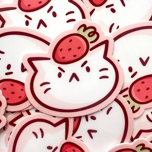 May include: A collection of stickers featuring a white cat face with a strawberry on its head. The strawberries are red with green stems, and the cat faces have a pink outline and angry expressions.