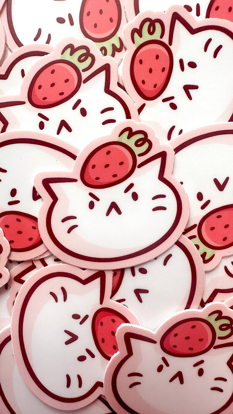 May include: A collection of stickers featuring a white cat face with a strawberry on its head. The strawberries are red with green stems, and the cat faces have a pink outline and angry expressions.