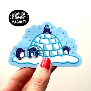 May include: A blue and white weatherproof magnet featuring four penguins huddled together in a snow-covered igloo. The text "WEATHER PROOF MAGNET!" is written in black on a white circle.