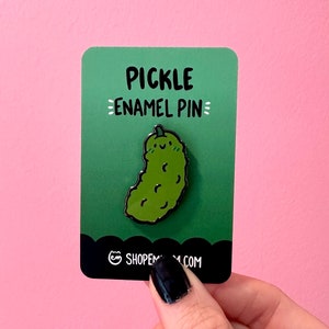 May include: Green enamel pin shaped like a pickle with a smiling face. The pin is on a green card with the text "Pickle Enamel Pin" and the website "shopmeow.com".