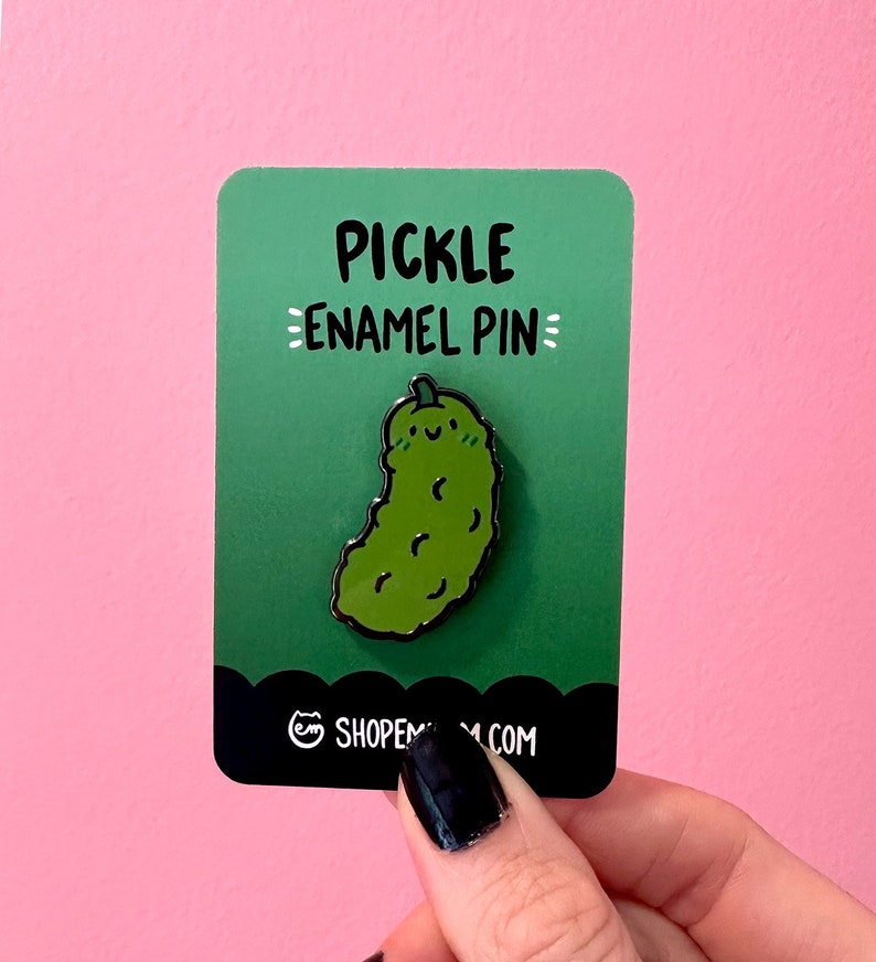 May include: Green enamel pin shaped like a pickle with a smiling face. The pin is on a green card with the text "Pickle Enamel Pin" and the website "shopmeow.com".