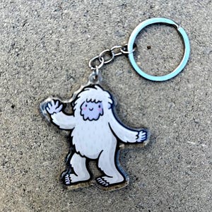 Puede incluir: Un llavero de acrílico transparente con un dibujo animado de un yeti con una cara azul claro y un llavero azul claro. El yeti es blanco con contornos negros y está en posición de caminar. El llavero está sujeto a una cadena plateada.