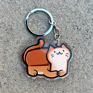 May include: A brown and white cartoon cat keychain with a silver key ring. The cat is sitting on its hind legs with its front paws crossed.