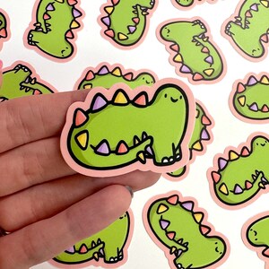 Op de afbeelding: Een set van 10 groene dinosaurus stickers met roze contouren en kleurrijke accenten. De dinosaurussen hebben vrolijke gezichten en staan in verschillende poses.
