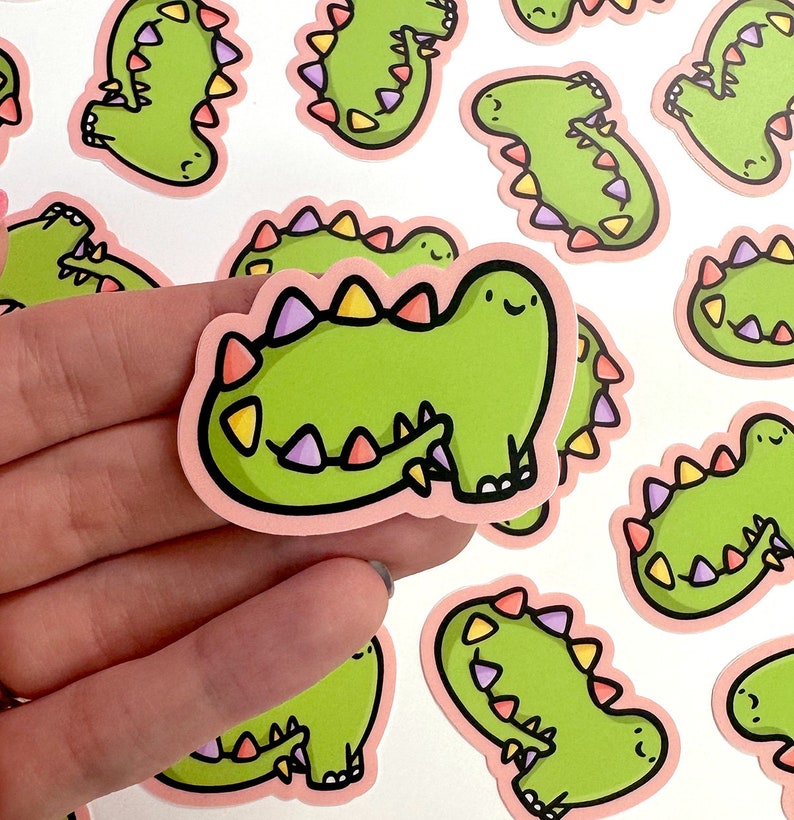 Op de afbeelding: Een set van 10 groene dinosaurus stickers met roze contouren en kleurrijke accenten. De dinosaurussen hebben vrolijke gezichten en staan in verschillende poses.