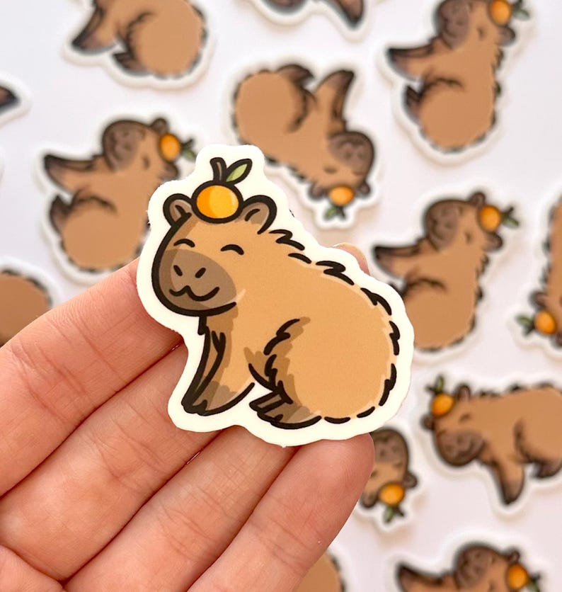 May include: A brown capybara sticker with a small orange on its head. The sticker is on a white background and is being held by a hand.