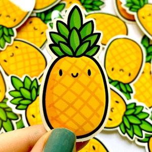 Op de afbeelding: Een verzameling gele ananasstickers met groene bladeren. Elke sticker heeft een lachend gezicht en een zwarte omtrek. De stickers staan op een witte achtergrond.