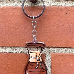 Op de afbeelding: Een transparante acryl sleutelhanger in de vorm van een koffiefilter met bruine koffie erin en een beige filter. De sleutelhanger heeft een zilveren ketting en ring.
