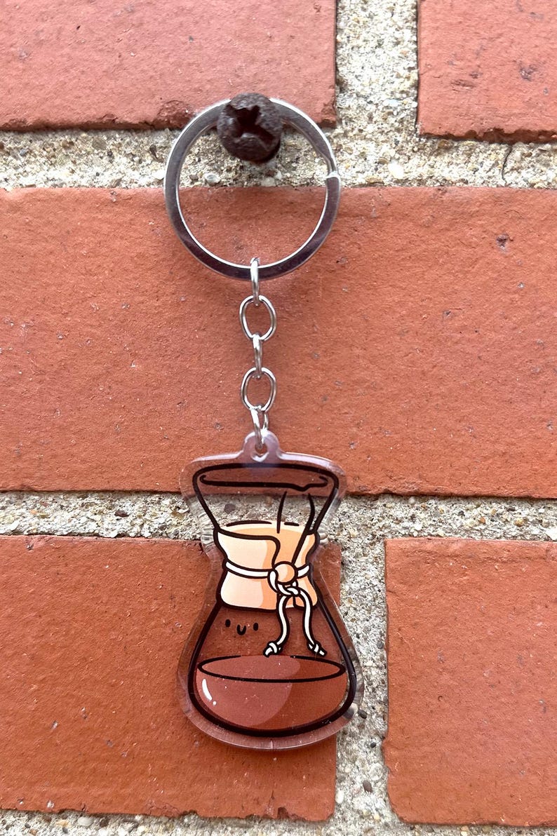 Op de afbeelding: Een transparante acryl sleutelhanger in de vorm van een koffiefilter met bruine koffie erin en een beige filter. De sleutelhanger heeft een zilveren ketting en ring.