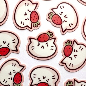 May include: A collection of stickers featuring a white cat with a strawberry. The cat has a simple, cartoon-like design with black facial features and a pink outline. The strawberries are red with green stems.