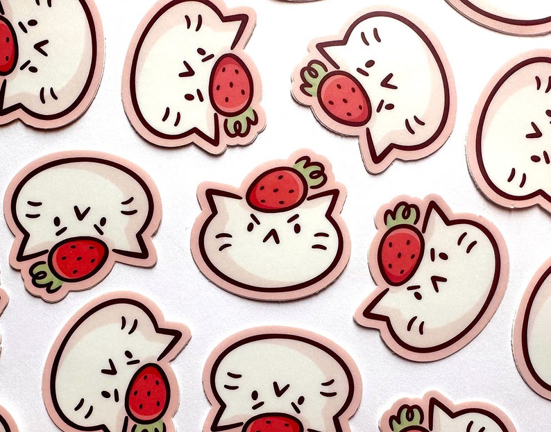May include: A collection of stickers featuring a white cat with a strawberry. The cat has a simple, cartoon-like design with black facial features and a pink outline. The strawberries are red with green stems.