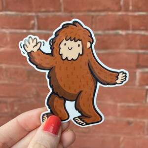 Puede incluir: Una pegatina de dibujos animados marr&oacute;n de un Bigfoot con una cara amigable, saludando con una mano. La pegatina est&aacute; sobre un fondo de ladrillo.