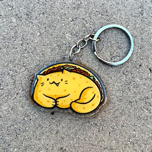 May include: A yellow cat-shaped keychain with a taco filling. The keychain is made of clear acrylic and has a silver metal key ring.