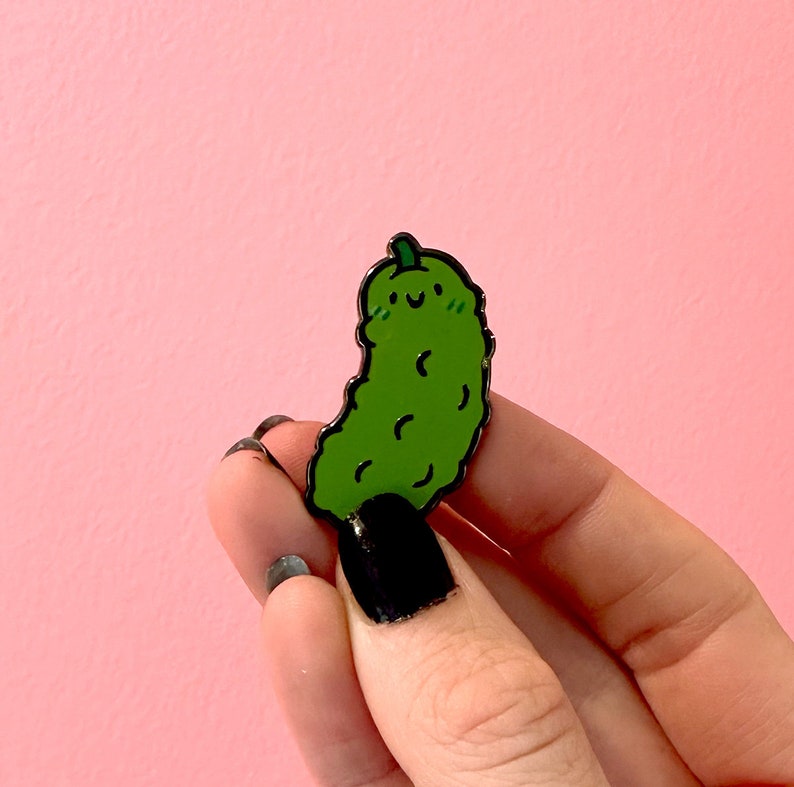 May include: A green pickle pin with a smiling face. The pin is made of metal and has a black outline.