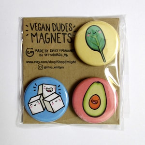 May include: Set of four round vegan-themed magnets. The magnets feature a smiling avocado, a smiling spinach leaf, and three smiling sugar cubes. The magnets are blue, yellow, pink, and light blue.