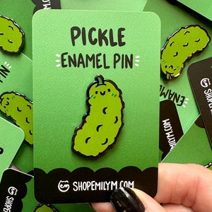 May include: A green enamel pin shaped like a pickle with a smiling face. The pin is on a green card with the text "PICKLE ENAMEL PIN" and the website "SHOPEMILYMM.COM".