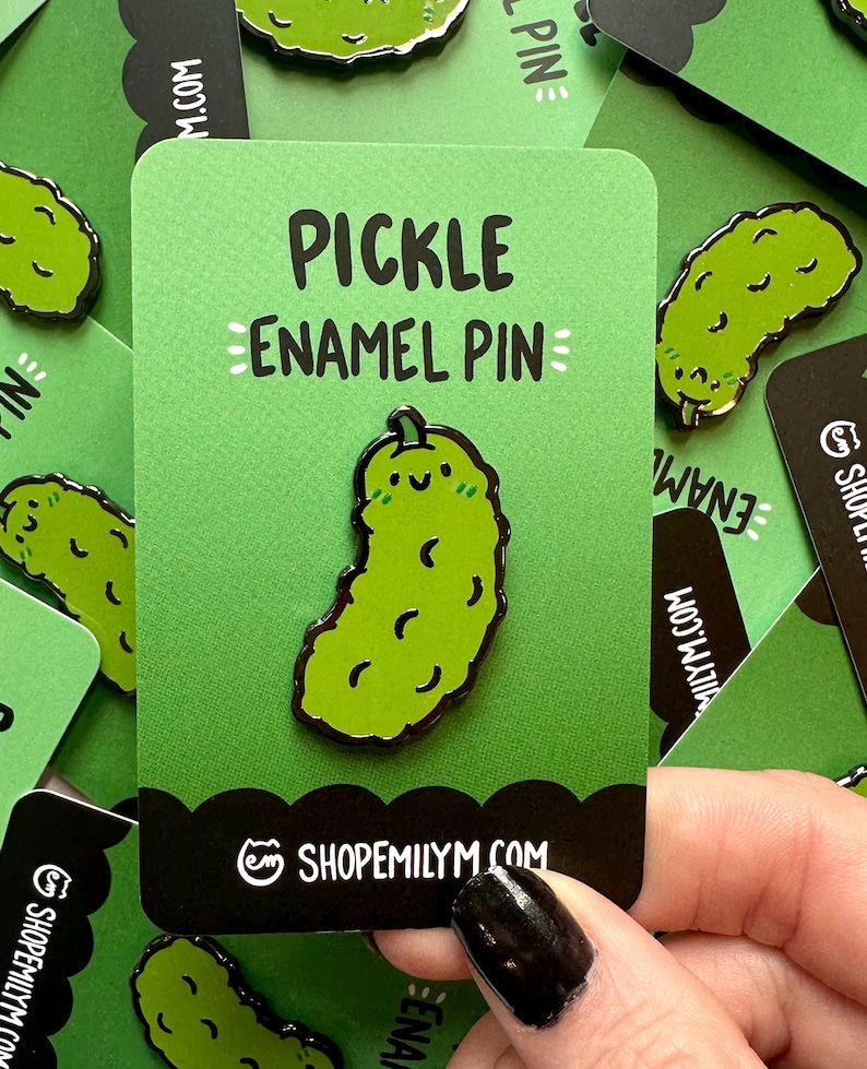 May include: A green enamel pin shaped like a pickle with a smiling face. The pin is on a green card with the text "PICKLE ENAMEL PIN" and the website "SHOPEMILYMM.COM".