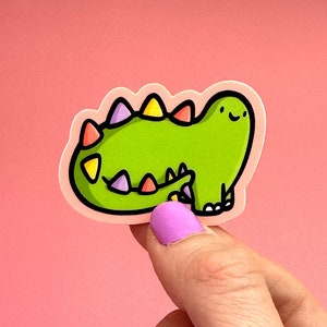 Op de afbeelding: Groene dinosaurus sticker met kleurrijke stekels en een glimlachend gezicht. De sticker is op een roze achtergrond.