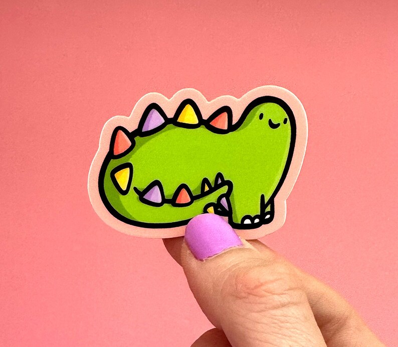 Op de afbeelding: Groene dinosaurus sticker met kleurrijke stekels en een glimlachend gezicht. De sticker is op een roze achtergrond.