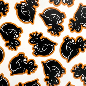 May include: A collection of black and orange stickers featuring a cartoon dragon wearing a bow tie. The dragon is in a variety of poses, including sitting, standing, and flying.