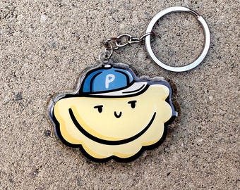 May include: A yellow cloud-shaped keyring with a smiling face and a blue baseball cap with a white brim and the letter "P". The keyring has a silver metal ring and chain.