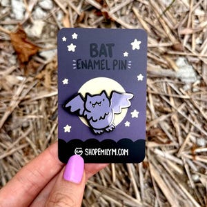 May include: Purple enamel pin of a cartoon bat with a yellow moon and white stars in the background. The text "BAT ENAMEL PIN" is written in black on the card. The card also has the website "SHOPEMILYMM.COM" written on it.